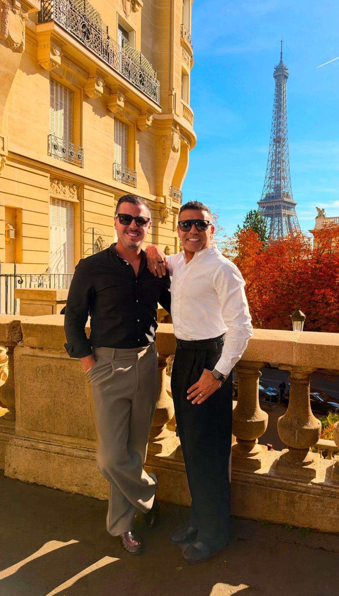 Elie and Justin on a stone balcony in Paris, the Eiffel Tower and autumn trees behind them